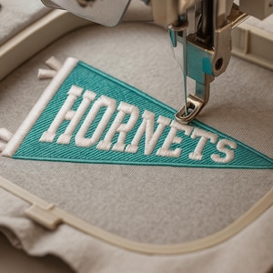 May include: Close-up of an embroidery machine stitching a teal pennant with the word "HORNETS" in white. The pennant is triangular with a white border and is being sewn onto a gray fabric. The machine's needle is visible.
