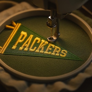 Può includere: Primo piano di una macchina da cucire che ricama un gagliardetto verde con la scritta "PACKERS" in filo d'oro. Il gagliardetto è in un telaio da ricamo. L'ago della macchina è posizionato sopra il tessuto, creando un design dettagliato e strutturato.