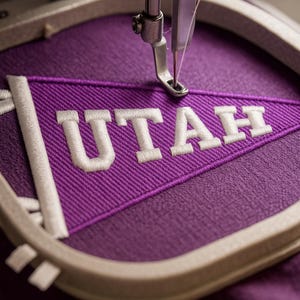 May include: Close-up of an embroidery machine stitching a purple pennant with the word "UTAH" in white thread. The pennant is held in a circular embroidery hoop. The needle is in the process of stitching the letter "A".