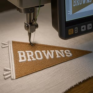 May include: An embroidery machine in action, creating a pennant. The pennant is a tan colour with the word "BROWNS" embroidered in white. The machine's screen shows the same design. The pennant is on white fabric.