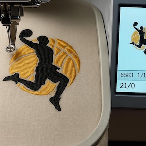 May include: An embroidered basketball design. It shows a black silhouette of a player dunking a ball, set against a yellow basketball. The design is on a beige fabric, suitable for a sports-themed project.