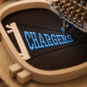 May include: Close-up of an embroidery machine stitching the word "CHARGERS" in blue thread onto black fabric. The machine's needle is actively working, creating the embroidered design within a white hoop. The image highlights the detail of the stitching process.
