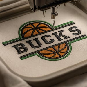 Puede incluir: Una máquina de bordar bordando un logotipo de "BUCKS". El diseño presenta letras negras, balones de baloncesto naranjas y bordes verdes y negros, todo sobre un fondo de tela blanca. La aguja de la máquina está enfocada.