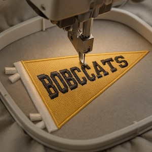 May include: A close-up of an embroidery machine stitching a yellow pennant with the text "BOBCATS" in black. The pennant has a white border and is set in a gray hoop. The machine needle is positioned over the fabric.