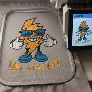 May include: Embroidered design featuring a smiling lightning bolt wearing sunglasses and giving a thumbs-up. The design is in shades of orange, blue, and black, with the text "LOS ANGELES" below. The design is on a gray fabric.