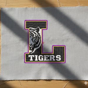 May include: Embroidered patch in the shape of the letter "L" with a tiger's face and the word "TIGERS". The patch has a black and white tiger design, outlined in purple and yellow, on a gray fabric background.