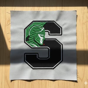 Op de afbeelding: Geborduurde patch met een gestileerde groene gladiatorenkop in een zwart-wit omrande letter 'S'. De patch staat op een lichtgrijze stoffen achtergrond. De gladiator heeft een wit gezicht en een rode stip op de kin.