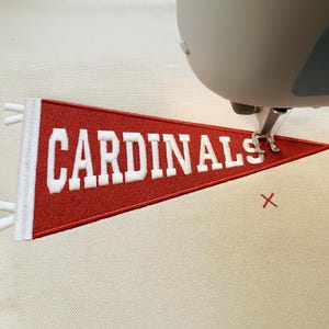 May include: A red pennant with the word "CARDINALS" embroidered in white. The pennant is being sewn by a machine, with the needle and presser foot visible. The background is a light beige fabric.