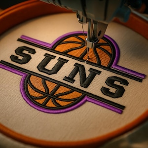 May include: Close-up of an embroidery machine stitching a Suns logo onto fabric. The logo features the word "SUNS" in black, with a basketball design in orange and black, and purple accents. The fabric is held in an orange hoop.