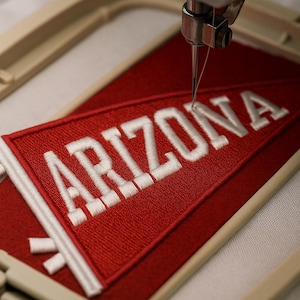 Op de afbeelding: Close-up van een borduurmachine die een rode wimpel met het woord "ARIZONA" in het wit borduurt. De wimpel zit in een beige borduurring, met de naald die actief de stof naait.