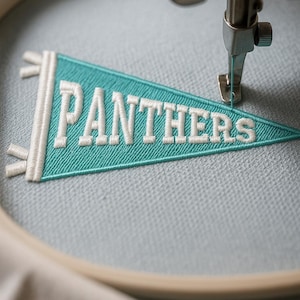 Pode incluir: Uma flâmula turquesa com a palavra "PANTHERS" bordada em branco, sendo costurada por uma máquina. A flâmula está em um bastidor de bordado de madeira, e o tecido é azul claro.
