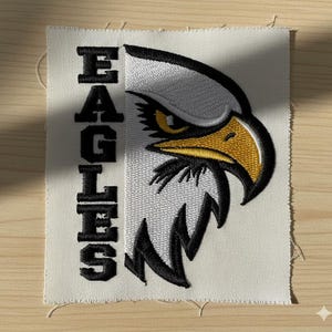 May include: Embroidered patch with an eagle head design. The eagle has a yellow beak and eye, and black and white feathers. The word "EAGLES" is vertically embroidered in black on the left. The patch is on a cream-coloured fabric.