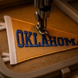 Könnte beinhalten: Nahaufnahme einer Stickmaschine, die einen goldenen Wimpel mit dem Wort "OKLAHOMA" in blauem Faden bestickt. Der Wimpel hat einen weißen Rand und wird auf einen braunen Stoff genäht.