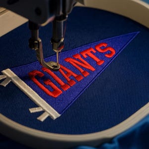 May include: A blue pennant with the word "GIANTS" embroidered in red thread is being sewn by a machine. The pennant has a white border and is held in a circular embroidery hoop.