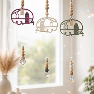 Könnte beinhalten: Drei hängende Ornamente mit hölzernen Wohnwagensilhouetten in Dunkellila, Beige und Olivgrün. Jeder Wohnwagen ist an einer Schnur mit Holzperlen und einem facettierten Kristallprisma am unteren Ende befestigt. Kleine rechteckige Anhänger sind angebracht.