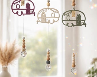 Puede incluir: Tres adornos colgantes con siluetas de caravanas de madera en color morado oscuro, beige y verde oliva. Cada caravana está suspendida por un hilo con cuentas de madera y un prisma de cristal facetado en la parte inferior. Incluyen etiquetas.
