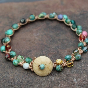May include: A handmade bracelet featuring turquoise, green, and multicolored beads strung on a beige cord. The bracelet includes a large, round, light-colored wooden bead as a focal point. The beads are various sizes and shapes.