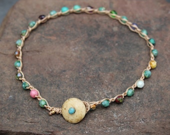 May include: Handmade necklace featuring a beige woven cord adorned with colorful beads in shades of turquoise, pink, yellow, and purple. A large, round, beige pendant with a turquoise center bead is the focal point. The necklace is displayed on a natural stone surface.