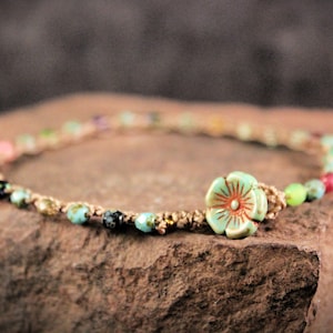 May include: A delicate ankle bracelet made with natural hemp cord and colorful beads, including turquoise, green, pink, and black. A ceramic flower charm in shades of blue and orange is the focal point of the bracelet.