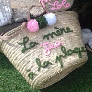 May include: Two straw tote bags with pom poms and embroidered text. The larger bag says "La mere Tea la plage" in green and pink yarn. The smaller bag says "Princesse Lola" in pink yarn.