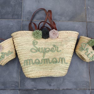 May include: Three straw tote bags with brown leather handles. The largest bag has green pom-poms and the text "Super Maman" embroidered in green. The smaller bags have green and white pom-poms and the text "Nova" and "Doris" embroidered in green.