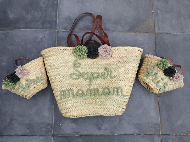 May include: Three straw tote bags with brown leather handles. The largest bag has green pom-poms and the text "Super Maman" embroidered in green. The smaller bags have green and white pom-poms and the text "Nova" and "Doris" embroidered in green.