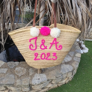 May include: A woven straw tote bag with brown leather handles and white and pink pom-poms. The bag is adorned with the letters "J & A" and the year "2023" in pink. This bag is a stylish accessory for carrying essentials, perfect for a day out.
