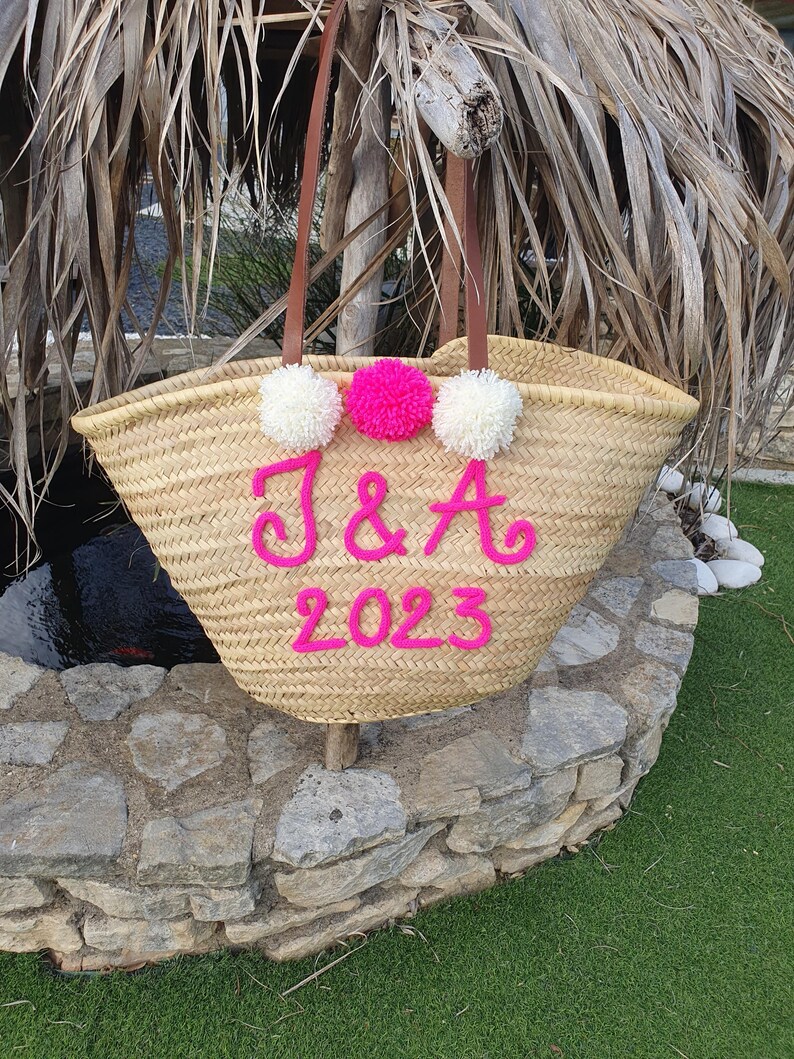 May include: A woven straw tote bag with brown leather handles and white and pink pom-poms. The bag is adorned with the letters "J & A" and the year "2023" in pink. This bag is a stylish accessory for carrying essentials, perfect for a day out.