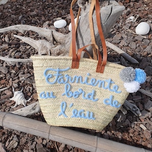 May include: Woven straw tote bag with brown leather handles. The bag features light blue embroidered text that reads "Farniente au bord de l'eau" and is decorated with grey, white, and blue pom-poms. The bag is designed for carrying items.