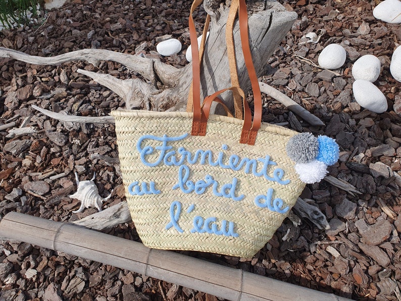 May include: Woven straw tote bag with brown leather handles. The bag features light blue embroidered text that reads "Farniente au bord de l'eau" and is decorated with grey, white, and blue pom-poms. The bag is designed for carrying items.