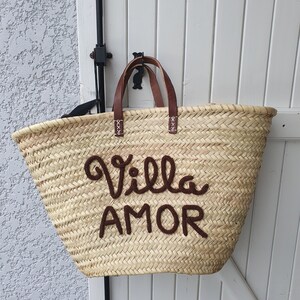 May include: Woven straw tote bag with brown leather handles and the words "Villa AMOR" embroidered in brown. The bag is hanging on a white wooden door. The bag is a light tan colour.