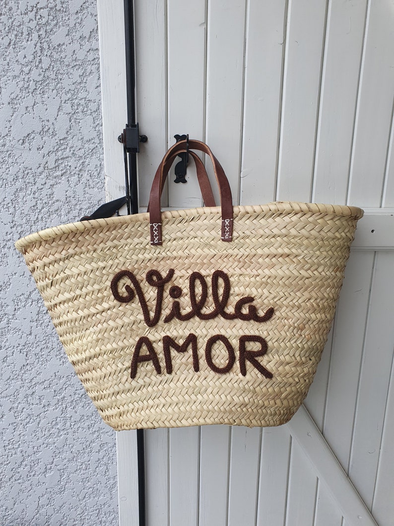 May include: Woven straw tote bag with brown leather handles and the words "Villa AMOR" embroidered in brown. The bag is hanging on a white wooden door. The bag is a light tan colour.