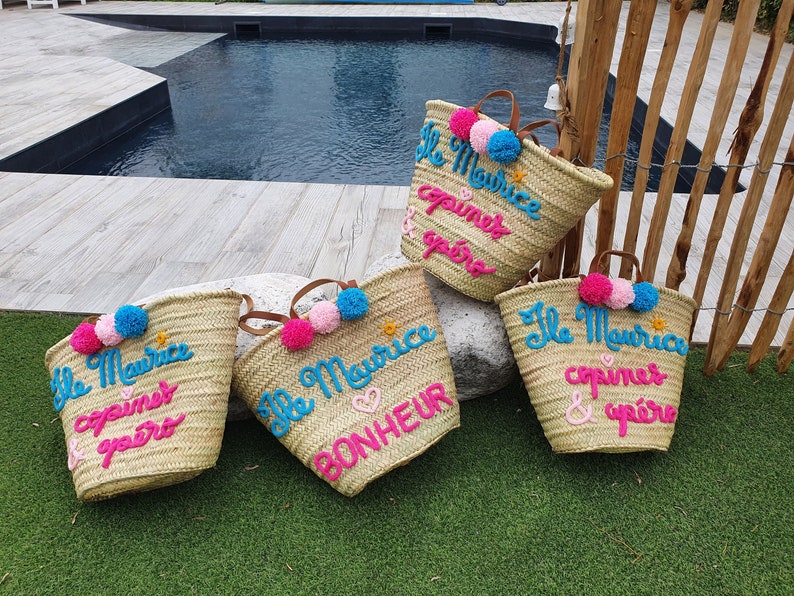 May include: Four straw tote bags with colorful pom-poms and embroidered text. The bags say "Ile Maurice" and "copines & ap&eacute;ro" in pink and blue lettering.