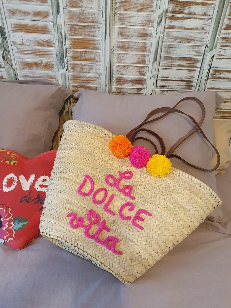 May include: A woven straw tote bag with brown leather handles and the phrase "La Dolce Vita" in pink script. The bag is decorated with three colourful pom-poms. It rests on a bed with a heart-shaped pillow.