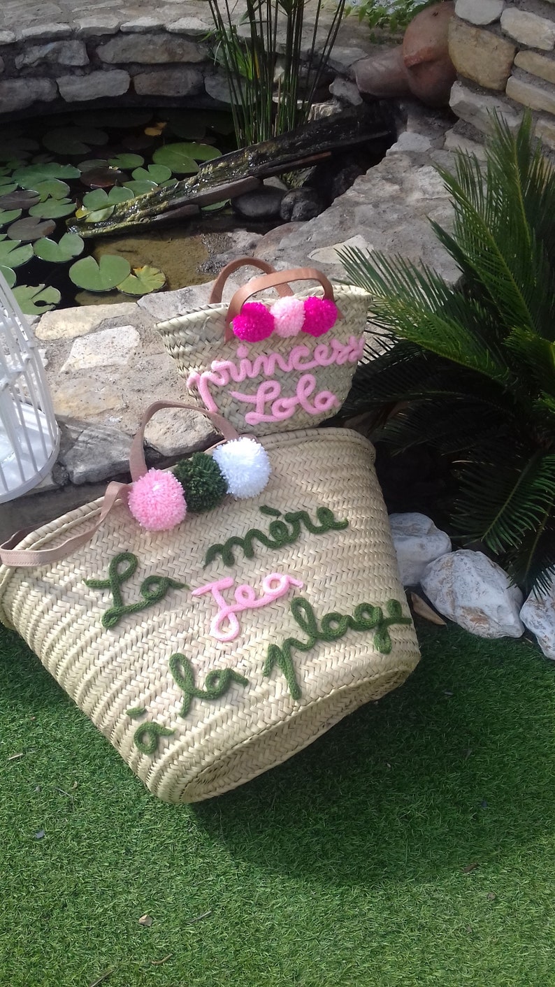 May include: Two straw tote bags with pom poms and embroidered text. The larger bag says "La mere Tea la plage" in green and pink yarn. The smaller bag says "Princesse Lola" in pink yarn.