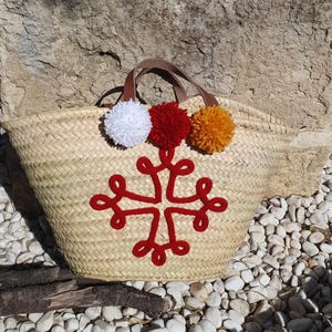 May include: Woven straw tote bag with brown leather handles and a red embroidered cross design. The bag is decorated with three colourful pom-poms: white, red, and orange. The bag is sitting on a bed of white pebbles.