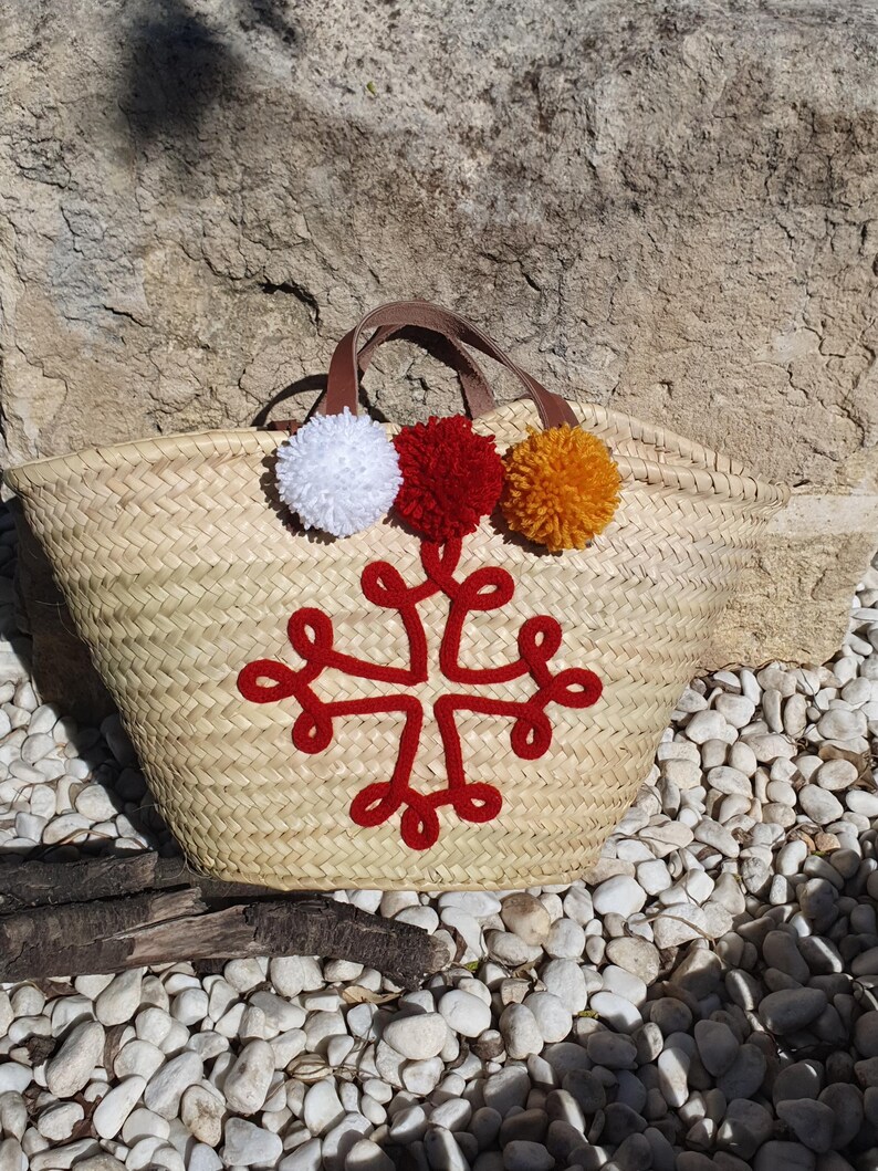 May include: Woven straw tote bag with brown leather handles and a red embroidered cross design. The bag is decorated with three colourful pom-poms: white, red, and orange. The bag is sitting on a bed of white pebbles.