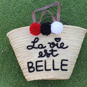 May include: Woven straw tote bag with brown leather handles and three pom-poms in red, black, and white. The words "La vie est BELLE" are embroidered in black on the front of the bag. The bag is sitting on green grass.