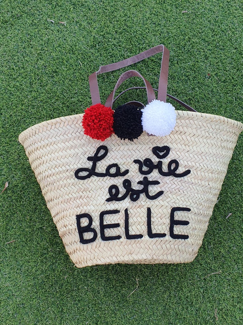 May include: Woven straw tote bag with brown leather handles and three pom-poms in red, black, and white. The words "La vie est BELLE" are embroidered in black on the front of the bag. The bag is sitting on green grass.