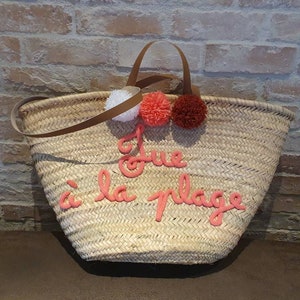 May include: A straw tote bag with a brown leather handle and pink pom-poms. The bag has the phrase "J'ue &agrave; la plage" embroidered on the front in pink.