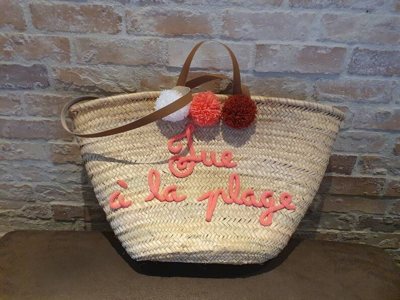 May include: A straw tote bag with a brown leather handle and pink pom-poms. The bag has the phrase "J'ue &agrave; la plage" embroidered on the front in pink.