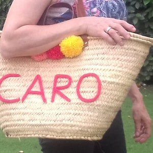 May include: Woven straw tote bag with the name "CARO" embroidered in red. The bag is decorated with colourful pom-poms and has a leather handle. The bag is being held in front of a green background.
