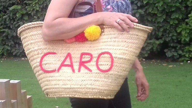 May include: Woven straw tote bag with the name "CARO" embroidered in red. The bag is decorated with colourful pom-poms and has a leather handle. The bag is being held in front of a green background.