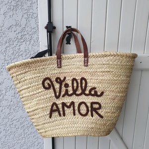 May include: A large, straw tote bag with brown leather handles. The bag has the words "Villa Amor" embroidered on the front in brown thread.