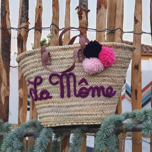 May include: Woven straw tote bag with brown leather handles and purple text that reads "La M&ocirc;me". The bag is decorated with three pom-poms in black, pink, and light pink. The bag is sitting in front of a wooden fence.