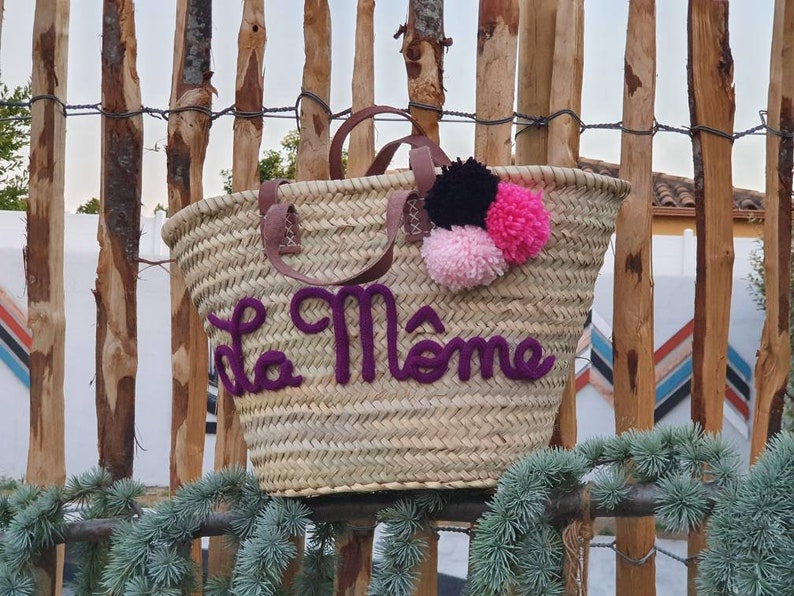 May include: Woven straw tote bag with brown leather handles and purple text that reads "La M&ocirc;me". The bag is decorated with three pom-poms in black, pink, and light pink. The bag is sitting in front of a wooden fence.