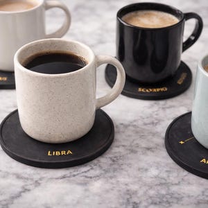 Könnte beinhalten: Vier Kaffeetassen auf schwarzen Untersetzern mit goldenen Tierkreiszeichen. Die Tassen sind weiß, schwarz und gesprenkelt beige, gefüllt mit Kaffee und Latte. Die Untersetzer haben die Tierkreiszeichen: Waage, Skorpion, Stier und Widder.
