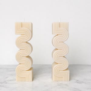 Peut inclure: Deux bougies sculpturales blanc cassé avec un design en zigzag unique. Chaque bougie a une base et un sommet carrés, avec un motif ondulé et superposé entre les deux. Les bougies sont non parfumées et ont une esthétique minimaliste.