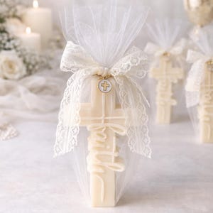 May include: Cream-coloured cross-shaped favours, each wrapped in sheer white fabric and tied with a lace-trimmed bow. The word "Faith" is written on the cross. A small charm with a cross is attached to the bow. Candles and flowers are in the background.