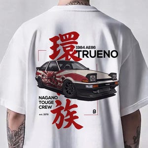 Könnte beinhalten: Ein weißes T-Shirt mit einer Grafik eines 1984 AE86 Trueno Autos mit roten und schwarzen Akzenten. Das Design enthält japanische Schriftzeichen sowie den Text "NAGANO TOUGE CREW est. 1979" und "1984 AE86 TRUENO" in Schwarz und Rot auf dem Rücken.